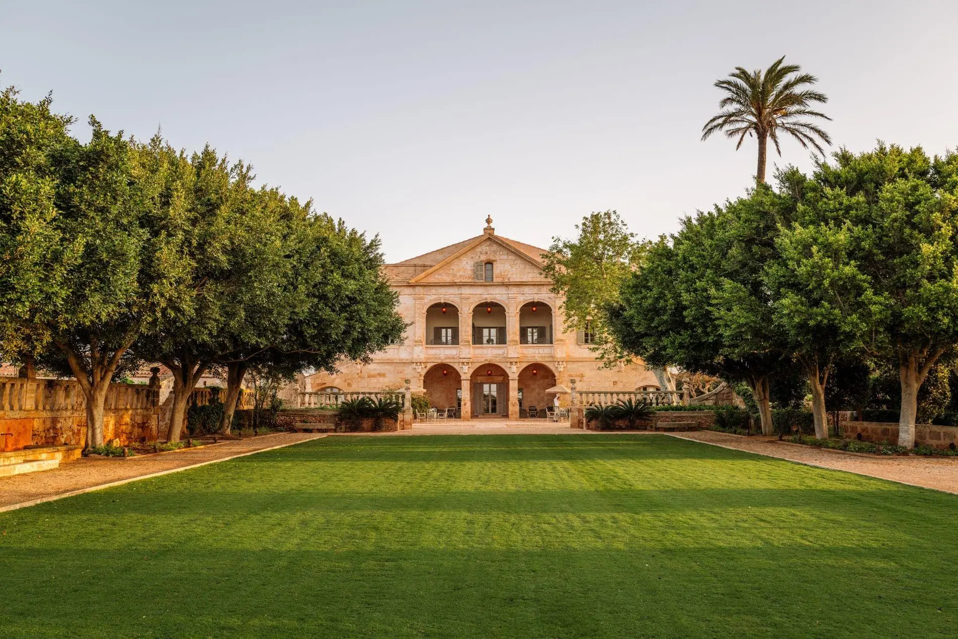 A historic stone estate house seen across its formal lawn and mature gardens at golden hour